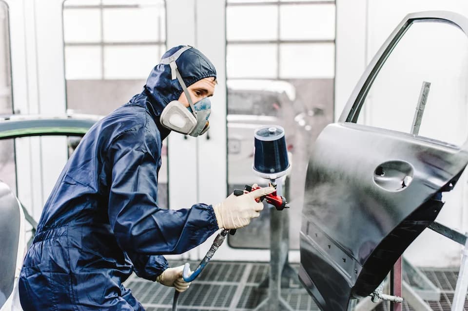 Lackierer in Schutzanzug und Atemschutz lackiert eine Fahrzeugtür in der Lackierkabine